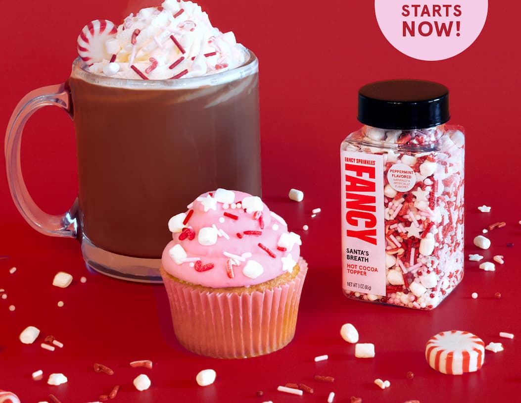 A glass mug of hot cocoa, a cute cupcake with pink frosting, both topped with Fancy Santa's Breath Hot Cocoa Topper sprinkles.