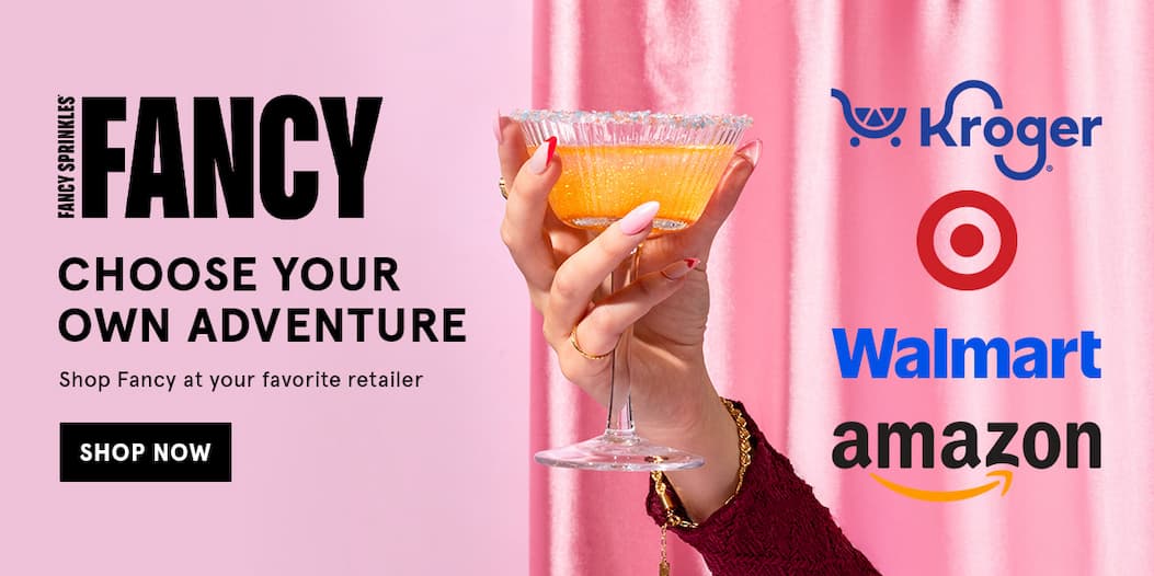 A hand holding a glass with a glittery cocktail is shown next to text that says Fancy Choose your own Adventure! Shop Fancy at your favorite retailer. Includes logos for Kroger, Target, Walmart, and Amazon. Click to shop now.