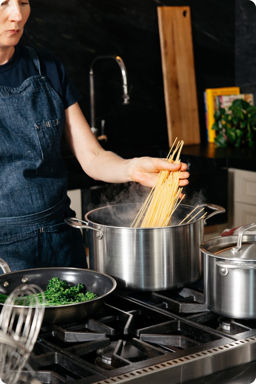 Cook adding pasta to a boiling stockpot with broccolini in a frying pan and a covered saucepan on a gas range