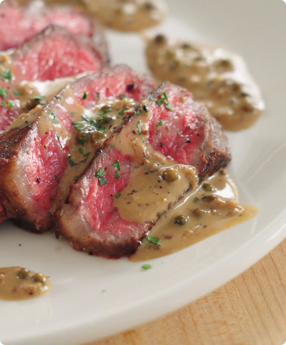 Sliced steak topped with peppercorn pan sauce on a white plate