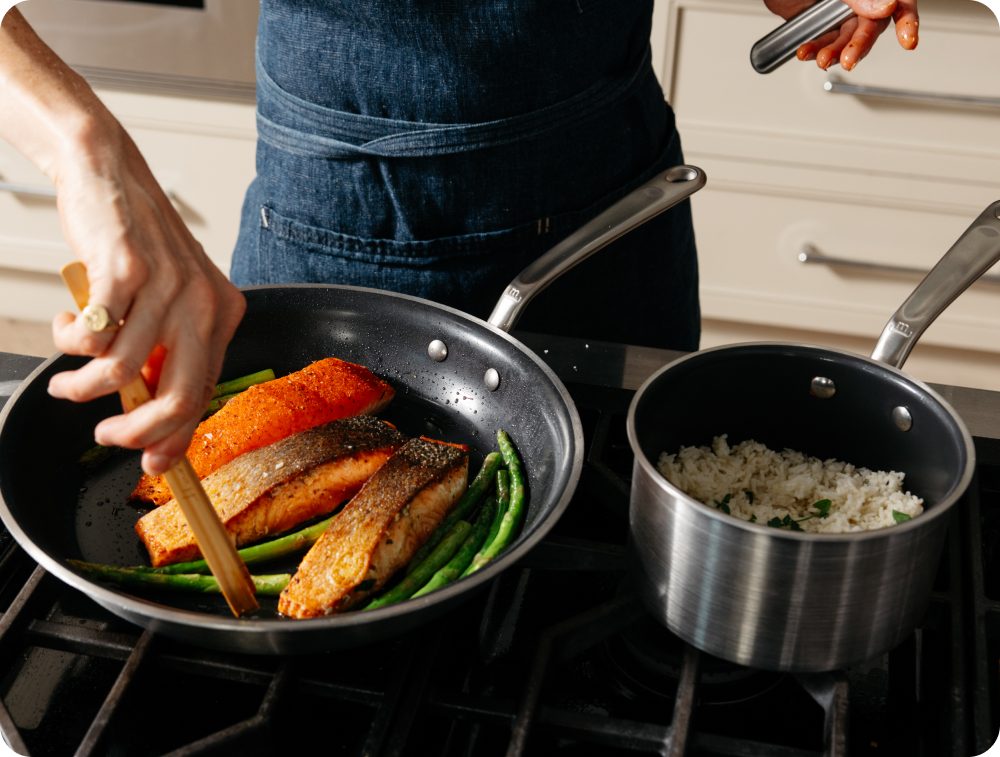 Cook plating crispy-skin salmon and green beans from a black CeramiClad frying pan next to a saucepan of rice on a gas stove