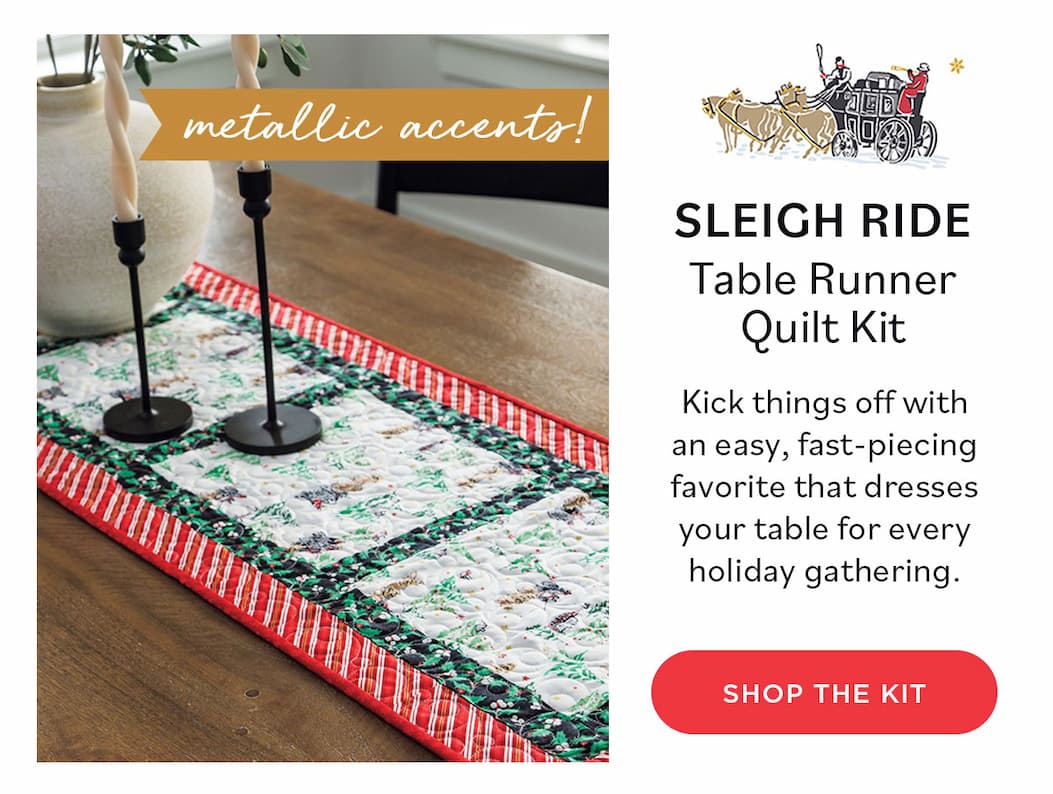 Image: A holiday themed quilted table runner in red, black, and white. Text: Metallic Accents! Sleigh Ride Table Runner Quilt Kit. Kick things off with an easy, fast-piecing favorite that dresses your table for every holiday gathering. Shop the Kit >>