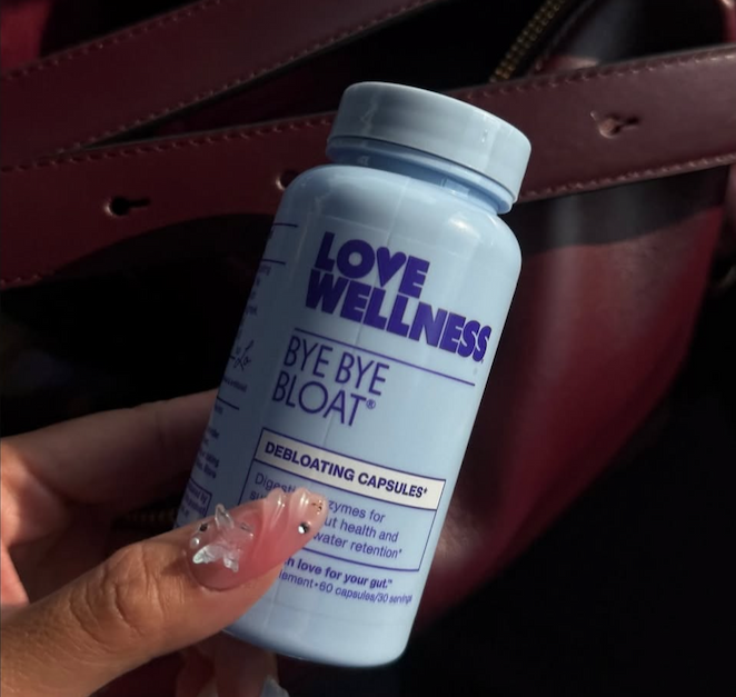 Hand holding a bottle of Love Wellness Bye Bye Bloat revitalizing capsules against a dark leather background.