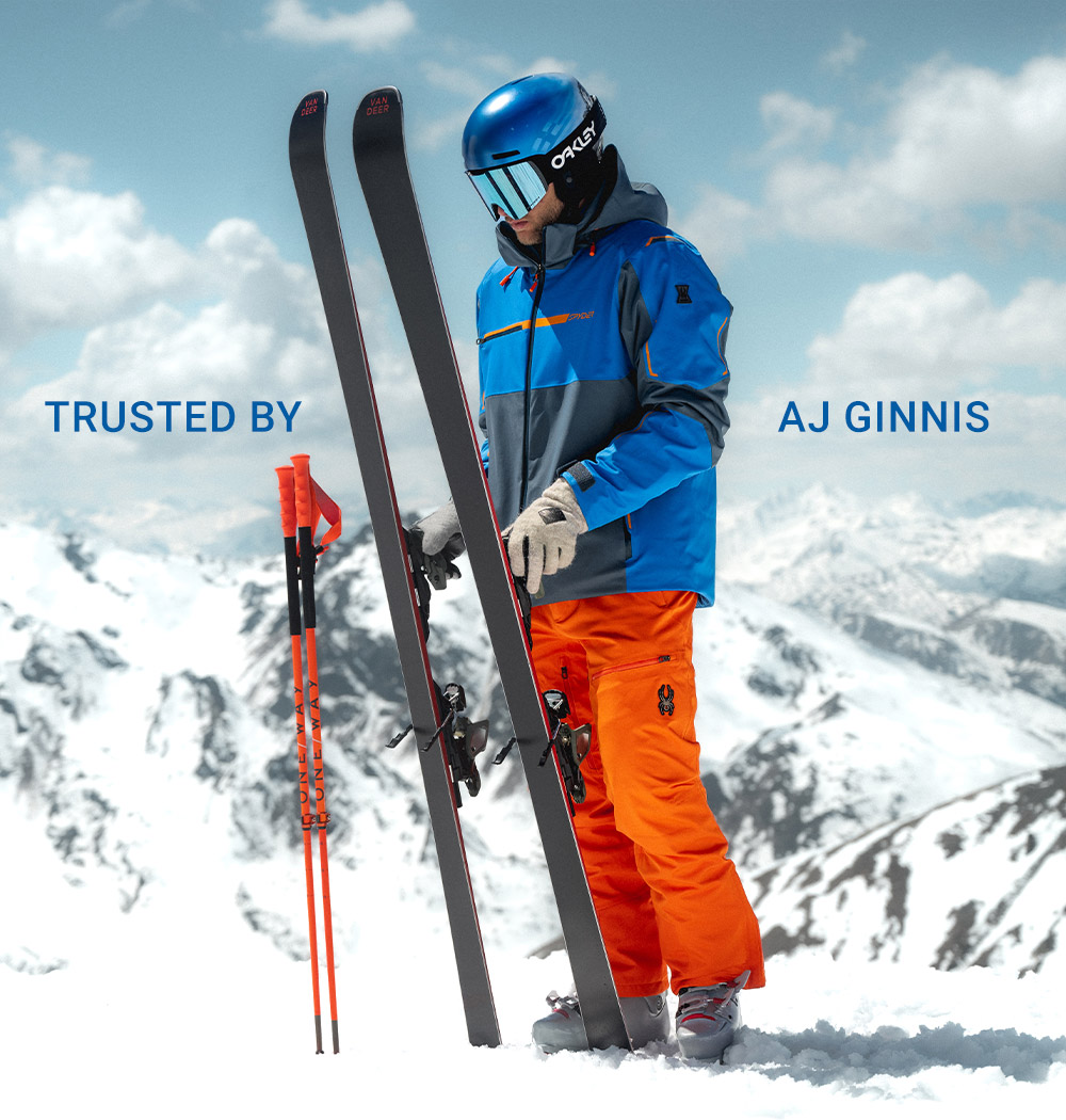 A skier in bright orange pants and a blue jacket holds skis and poles on a snowy mountain peak with dramatic mountain scenery.