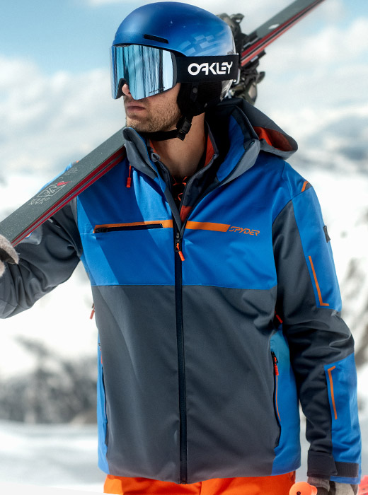 Skier standing against a snow background in a blue and grey ski jacket holding skis