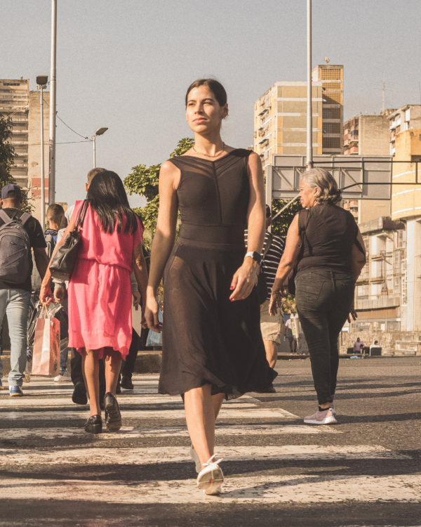 A woman in an elegant, sleeveless black summer dress walks confidently through a busy, sunny city street. In the background, other pedestrians are seen from behind, and the warm light creates a bright summer atmosphere.