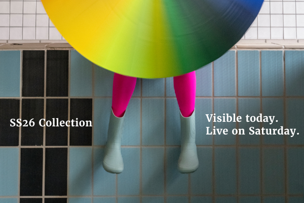 The image shows the legs of a person sitting on the edge of a swimming pool, their face and upper body covered by a rainbow-colored umbrella, wearing Ranidae mint. Next to the legs, “SS Collection 26” can be read on the left and “Visible today. Live on Saturday.” on the right.