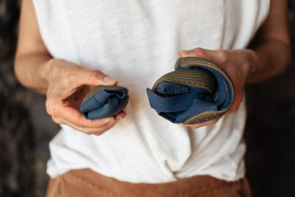 Close-up of two hands effortlessly rolling up a pair of dark blue Wildling shoes into two extremely small, compact bundles. This demonstrates the flexibility of the sole and how little space the shoes take up in luggage.