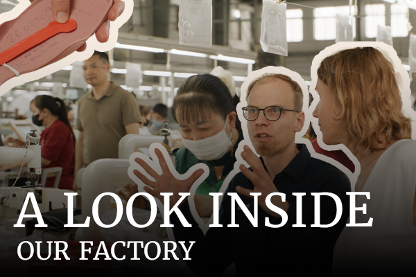 The image shows Anna Yona, founder of Wildling Shoes, talking to Marco, founder of Nordwolle. In the background, workers can be seen in a bright sewing factory. The image contains graphic elements: a Wildling sole outlined in white, and the text “A look inside our factory.”