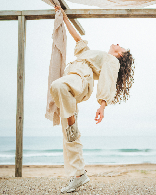 The picture shows a person on the beach. The sea can be seen in the background. The person is wearing beige summer clothes and the Lellin sablel. Above them is a pergola with a cloth stretched across it, which the person is holding onto.
