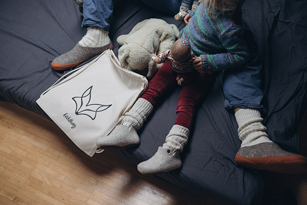 An adult and a child sitting on a sofa, wearing Slinka sten and Lotus slippers, with a Wildling tote bag beside them.