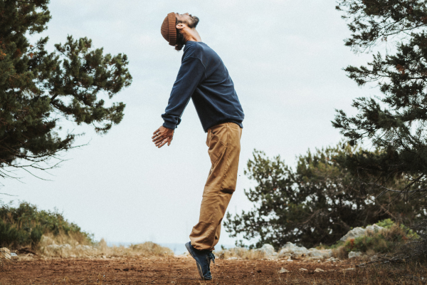  A man dances on a forested plateau, trees on either side. He stands on his toes, chest lifted, looking up at the sky. He wears a beanie, comfortable clothing, and Wildling Risaik.