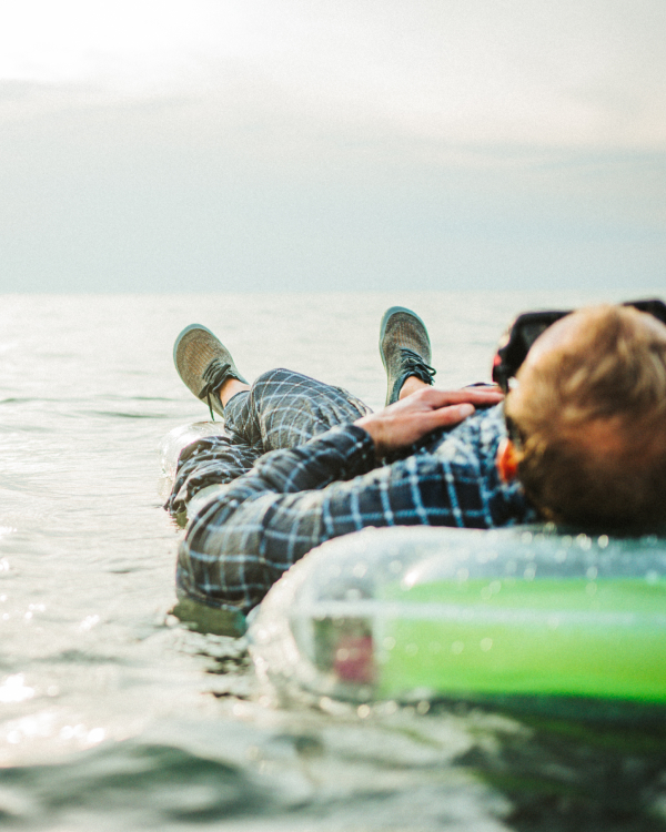 A person lies relaxed backwards on a green inflatable boat in the water, enjoying the view of the wide lake. They are wearing plaid trousers and green, flexible Wildling minimalist shoes.