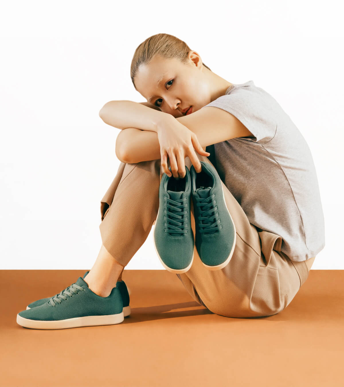 A person in casual attire sits on the floor, holding green sneakers. They wear beige pants and a gray T-shirt, exuding a calm, introspective vibe.