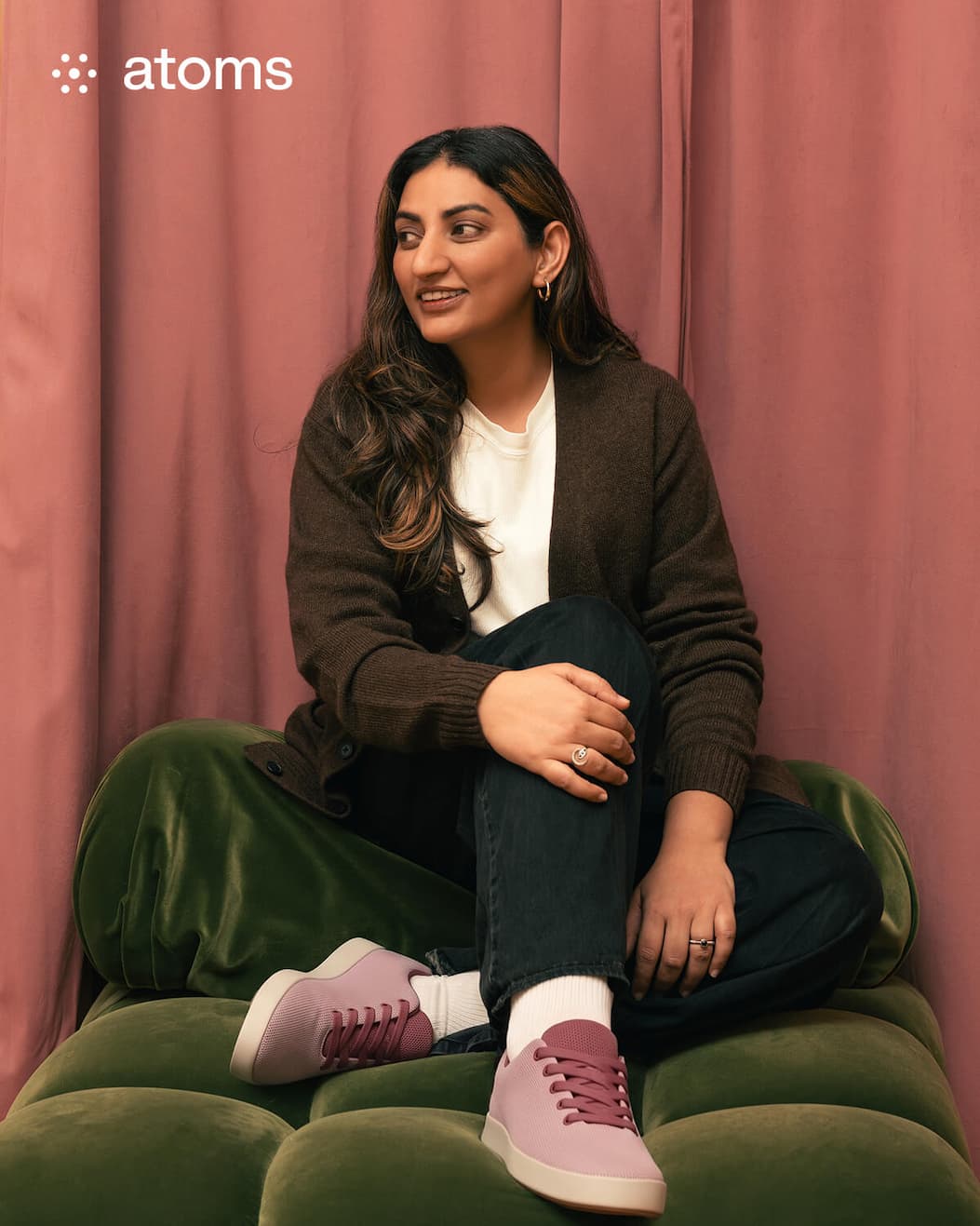 A person with long hair sits on a green couch against a mauve curtain, smiling and relaxed. They're wearing a brown cardigan, black pants, and pastel sneakers. The "atoms" logo is in the top left corner.