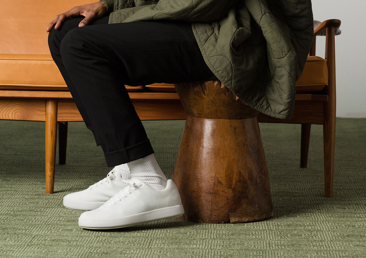 "Person in black pants and white sneakers sits on a wooden stool beside a tan couch, wearing a green jacket."