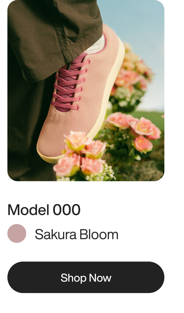 A pink sneaker with maroon laces steps on grass near vibrant pink flowers. The image is branded as "Model 000, Sakura Bloom." Tone is fresh and lively.