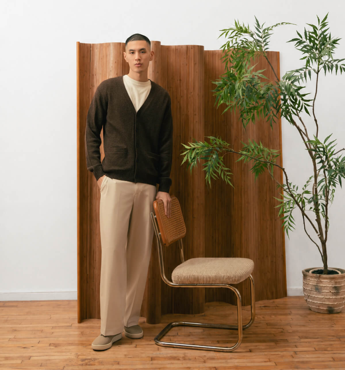 A man in a brown cardigan and beige pants stands beside a cane chair in a room with a potted plant and wooden panel backdrop, conveying a calm, minimalist vibe.