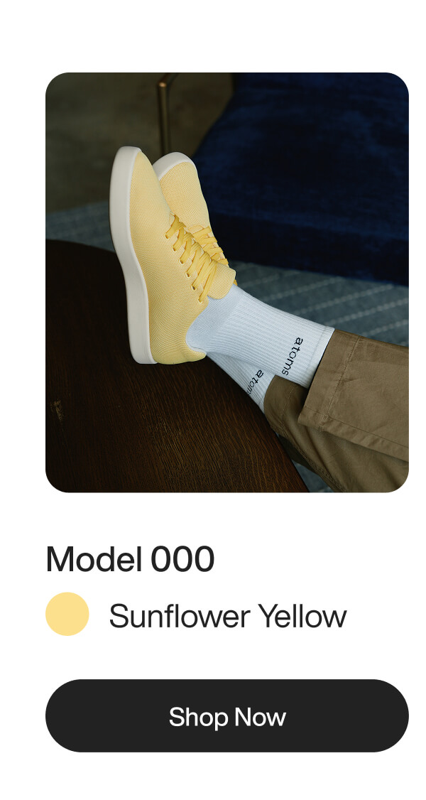 A person wearing sunflower yellow sneakers with white socks rests feet on a wooden table. Text below reads "Model 000, Sunflower Yellow, Shop Now."