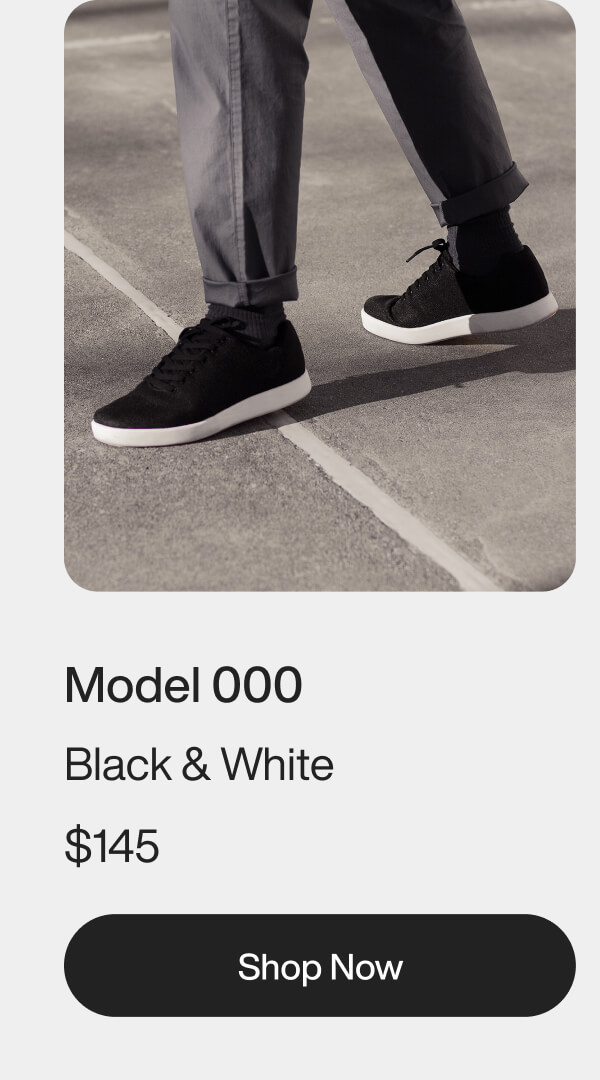 A person wearing gray pants and black sneakers with white soles stands on a pavement. Text below reads: "Model 000, Black & White, Shop Now."