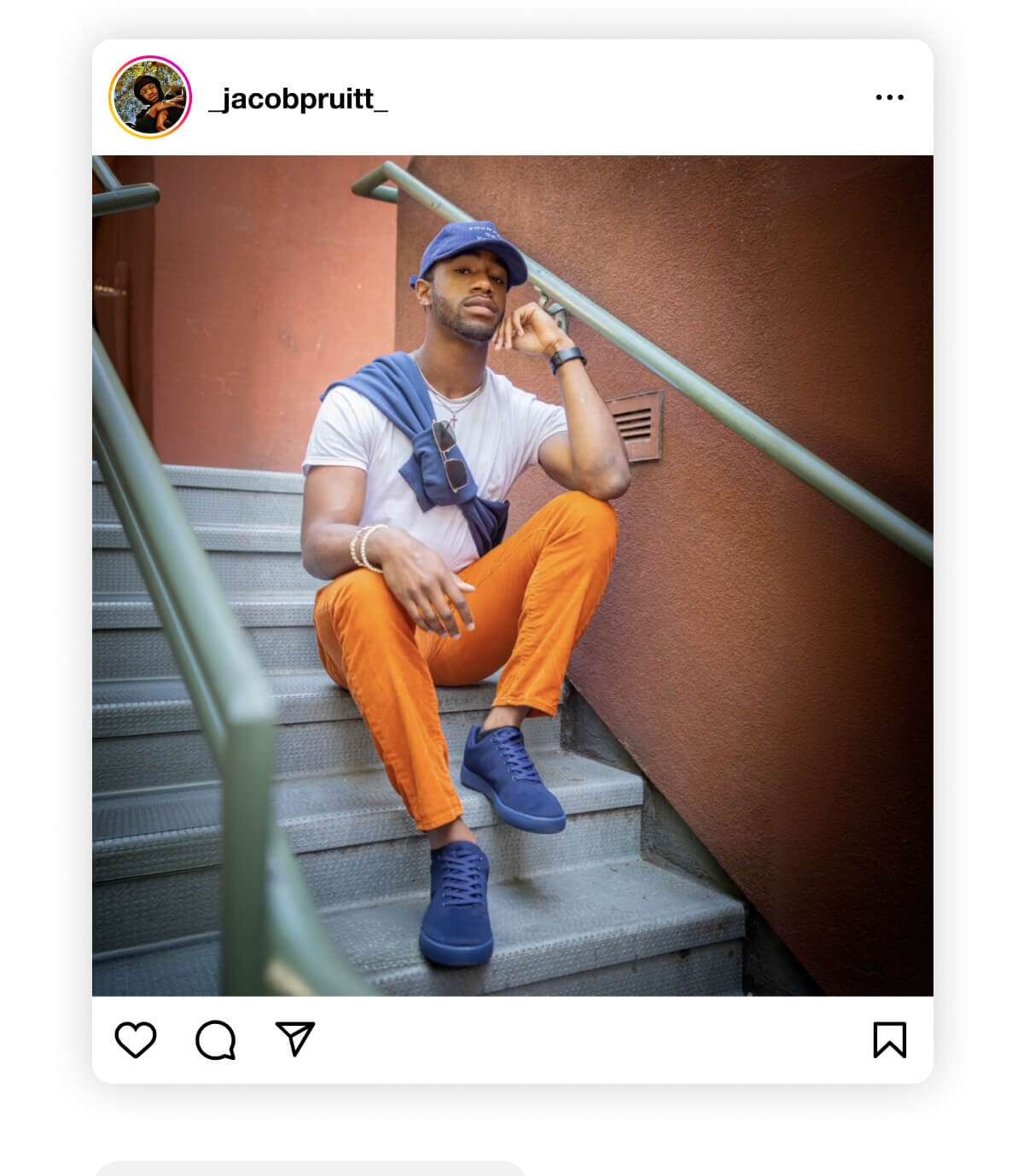 A man sits on outdoor stairs against a warm-toned wall, wearing a blue cap, white t-shirt, orange pants, and blue shoes, exuding a relaxed, stylish vibe.