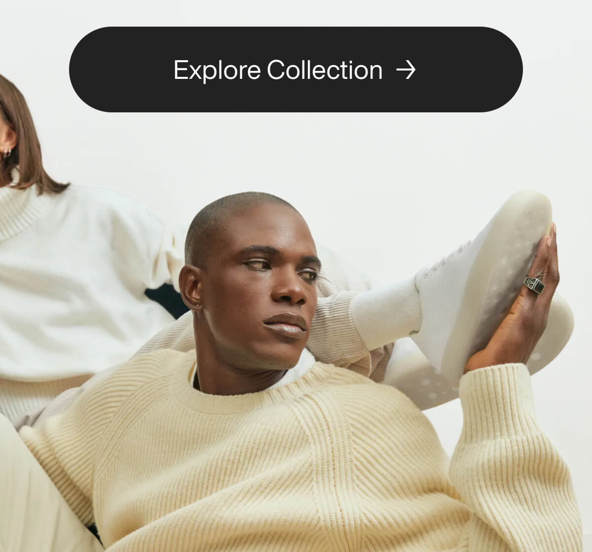 Man in a cream sweater reclines thoughtfully, holding a white sneaker. A woman in similar attire sits nearby. Text above reads "Explore Collection."