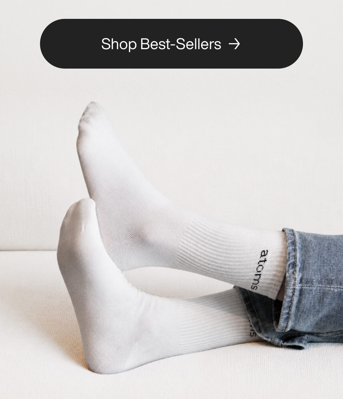 "White socks with "atoms" logo on crossed feet, denim jeans. "Shop Best-Sellers" button above. Minimalist and cozy vibe."