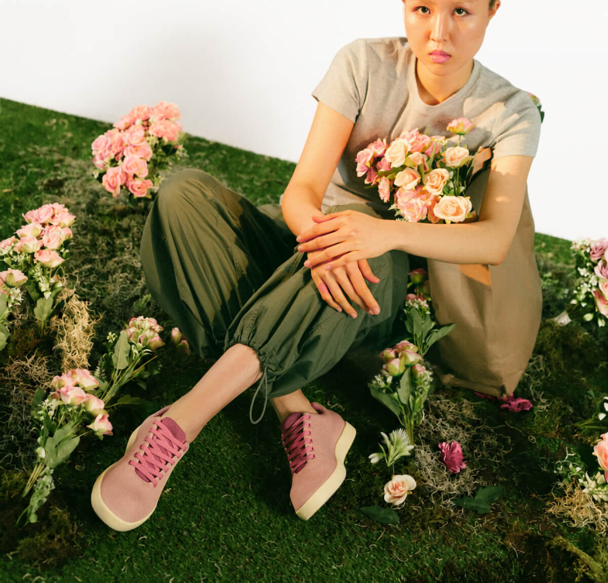 A person sits on grass surrounded by pink flowers, wearing a gray shirt, green pants, and purple shoes, holding a bouquet, conveying a calm, floral vibe.