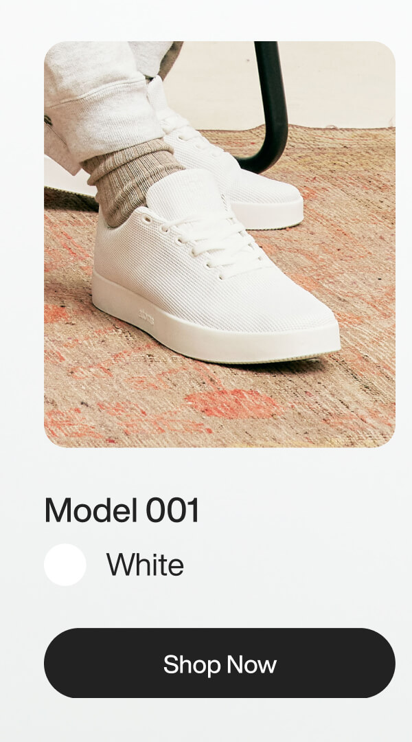 Close-up of white sneakers, "Model 001," on a textured rug. Worn with socks and sweatpants. Includes a "Shop Now" button below.