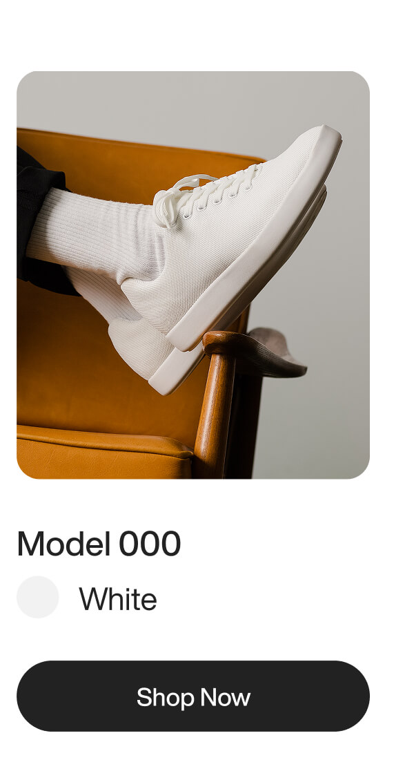 A minimalist image of a person wearing a sleek white sneaker with white socks. The foot is propped on a stylish orange chair. Text reads “Model 000, White.”