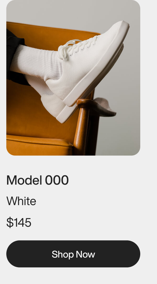 A white sneaker, worn with white socks, rests on an orange chair with wooden arms. Below are the words "Model 000 White" and a "Shop Now" button.