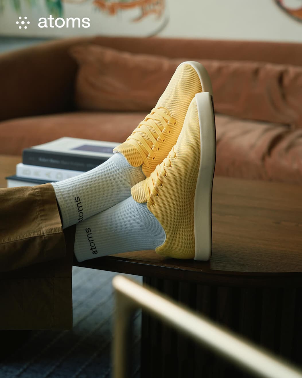 Yellow sneakers with white soles are propped on a table. The wearer has light blue socks branded with "atoms." Background: brown couch and books.