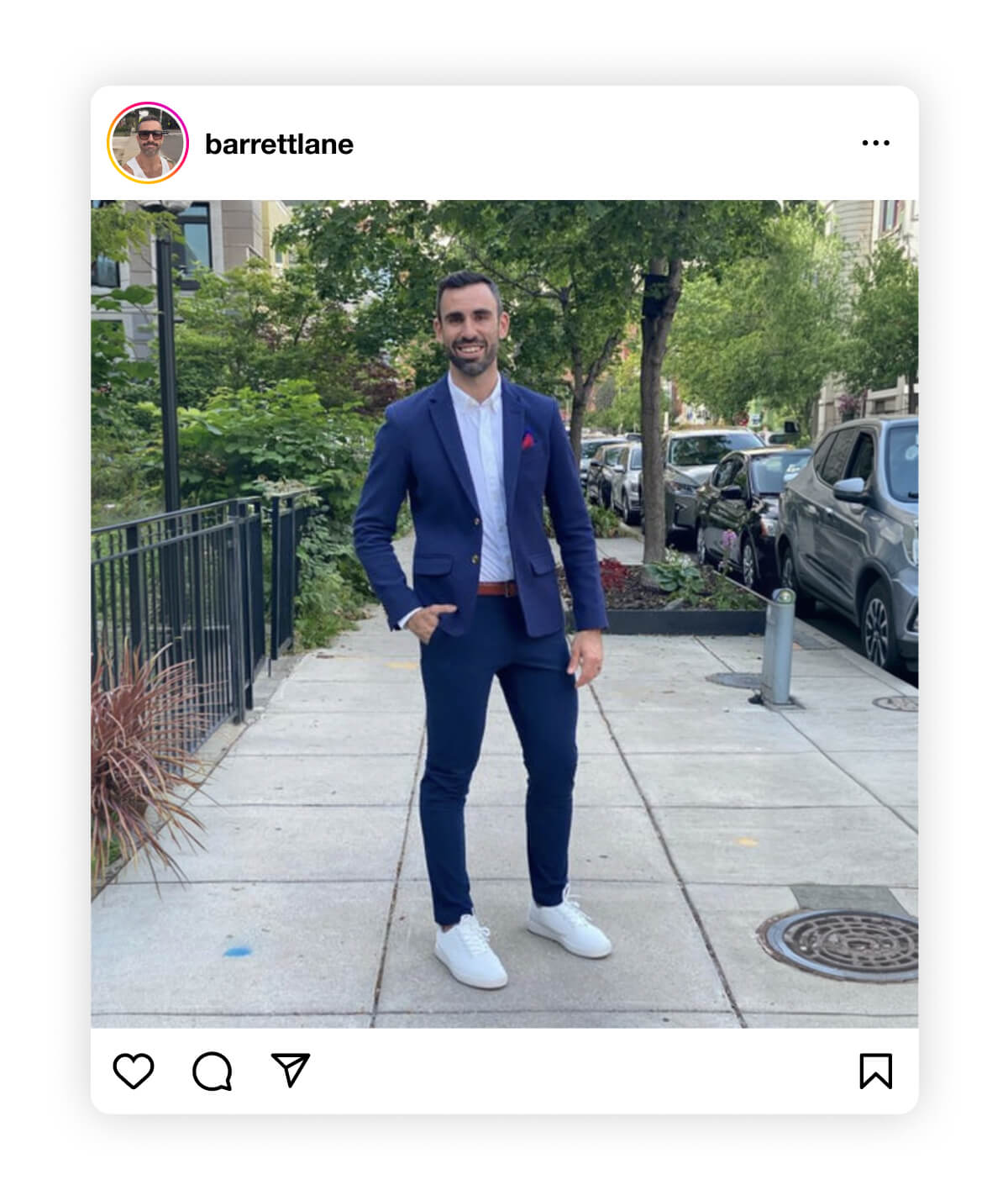Man in a navy suit and white sneakers stands smiling on an urban sidewalk, surrounded by trees and parked cars, conveying a relaxed, stylish vibe.
