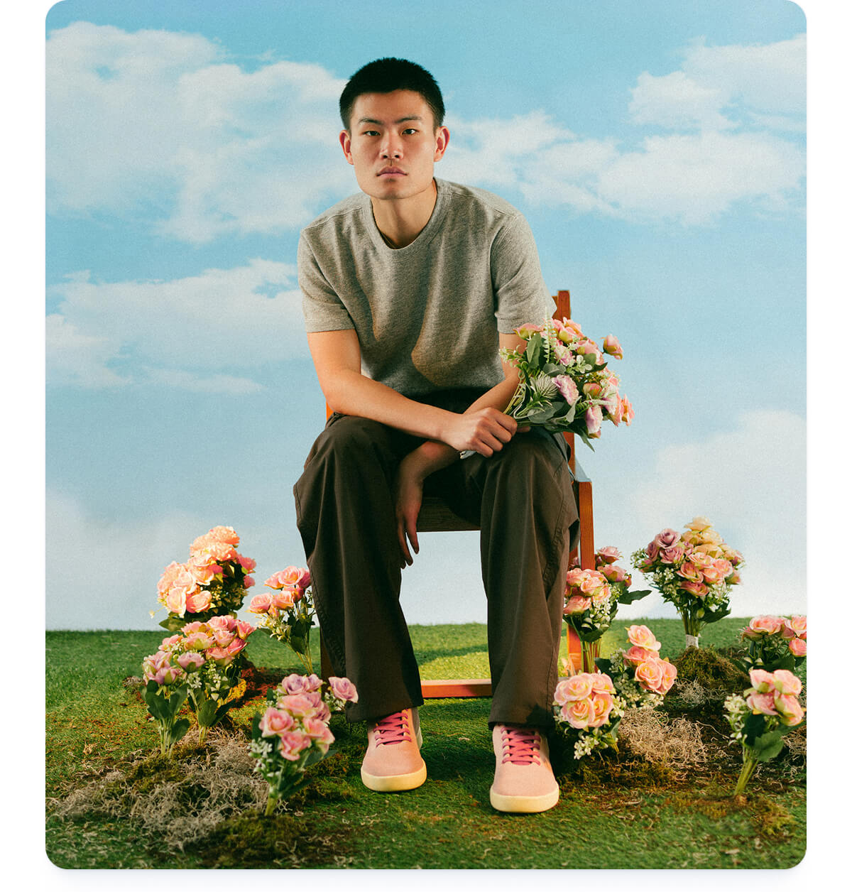 A person sits on a wooden chair holding flowers, surrounded by pink blooms on grassy terrain. The backdrop is a serene blue sky with clouds.