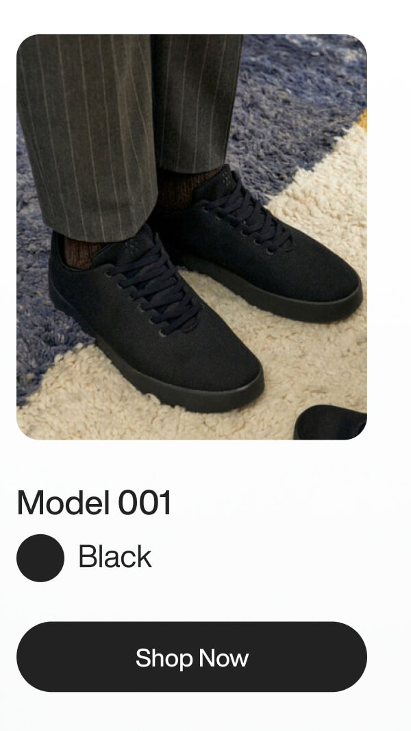 Black sneakers with black laces, worn with gray pinstriped pants, on a textured blue and beige carpet. Text: "Model 001, Black, Shop Now."