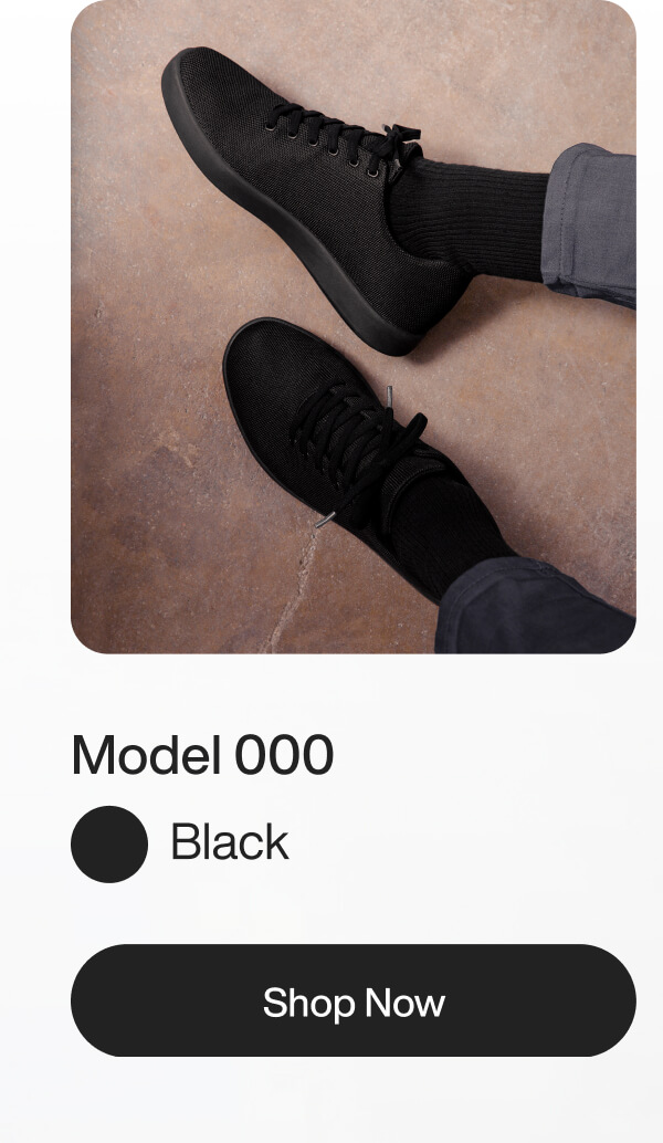 A person wearing black sneakers and socks sits on a tan floor. The sneakers are identified as "Model 000." A "Shop Now" button is visible below.