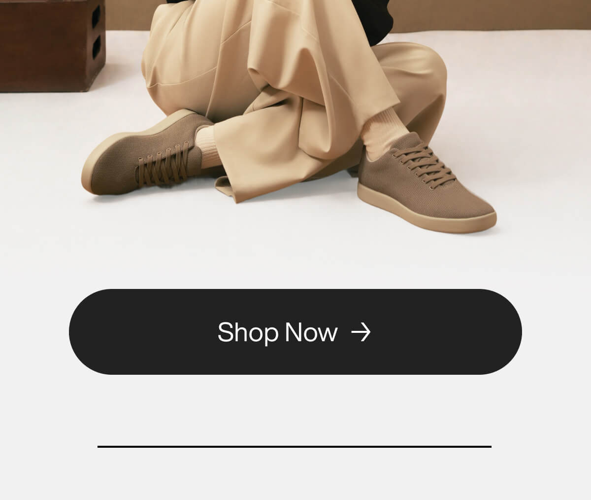 Person wearing beige trousers and brown sneakers sits on the floor against a minimal backdrop. A "Shop Now" button is at the bottom center.
