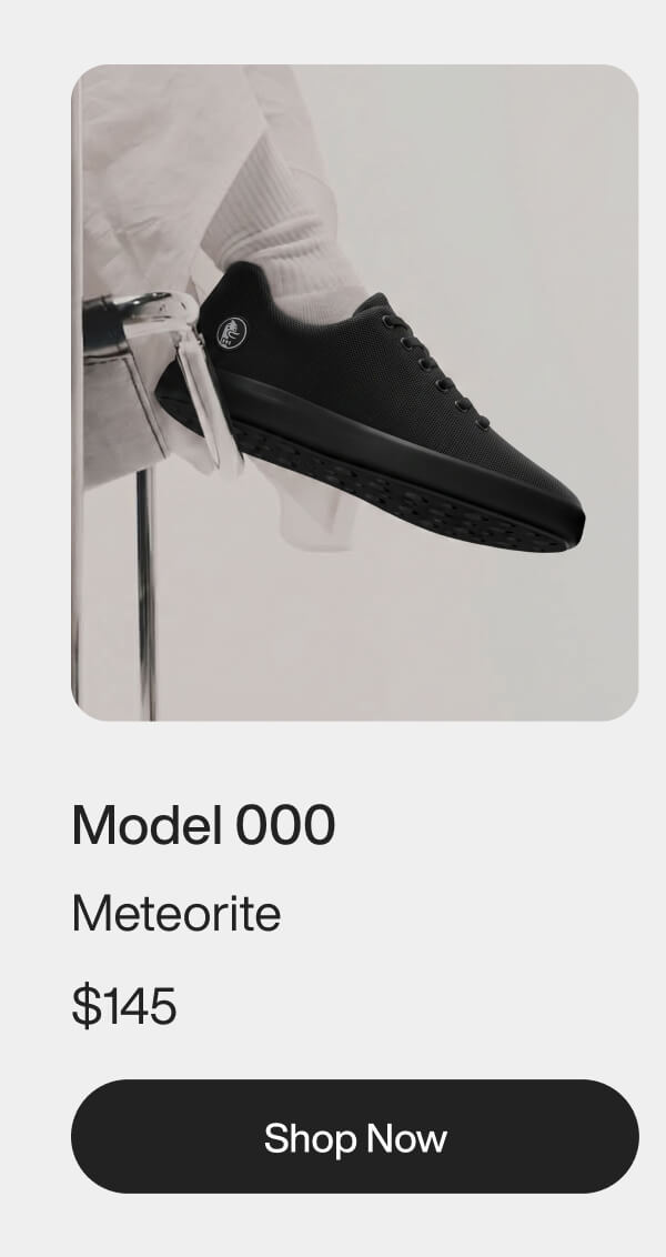 Black sneaker with a sleek design, labeled "Model 000 Meteorite," is worn by a person in white pants, perched on a stool. Modern and stylish tone.