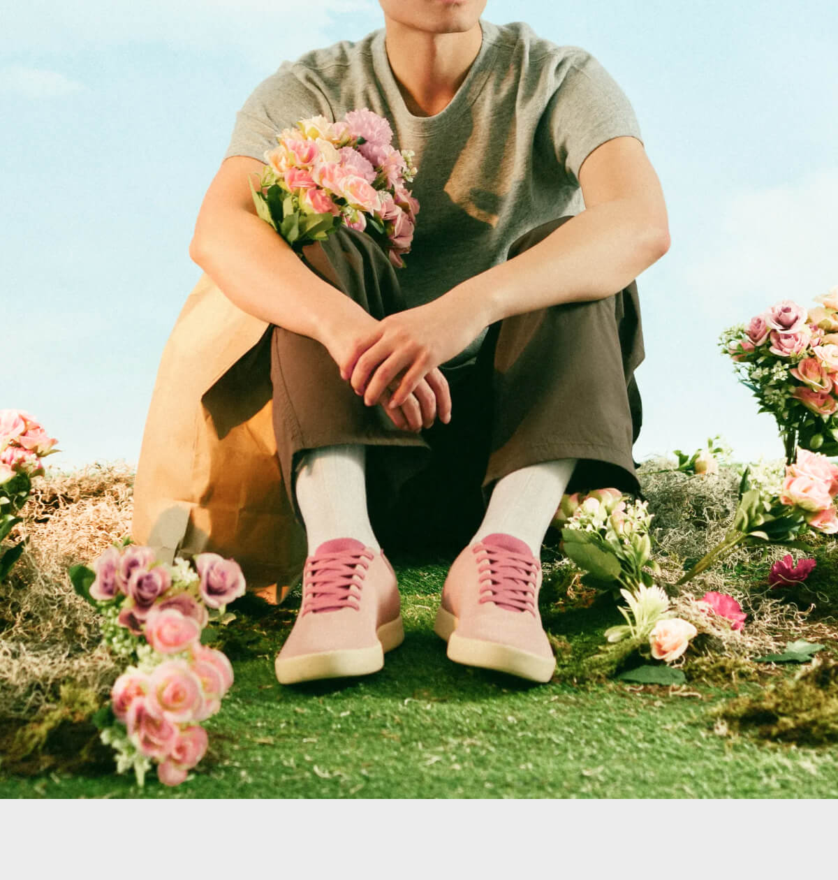 "Person in gray shirt and brown pants sits on grass, holding pink flowers. Pink sneakers and more flowers surround them."