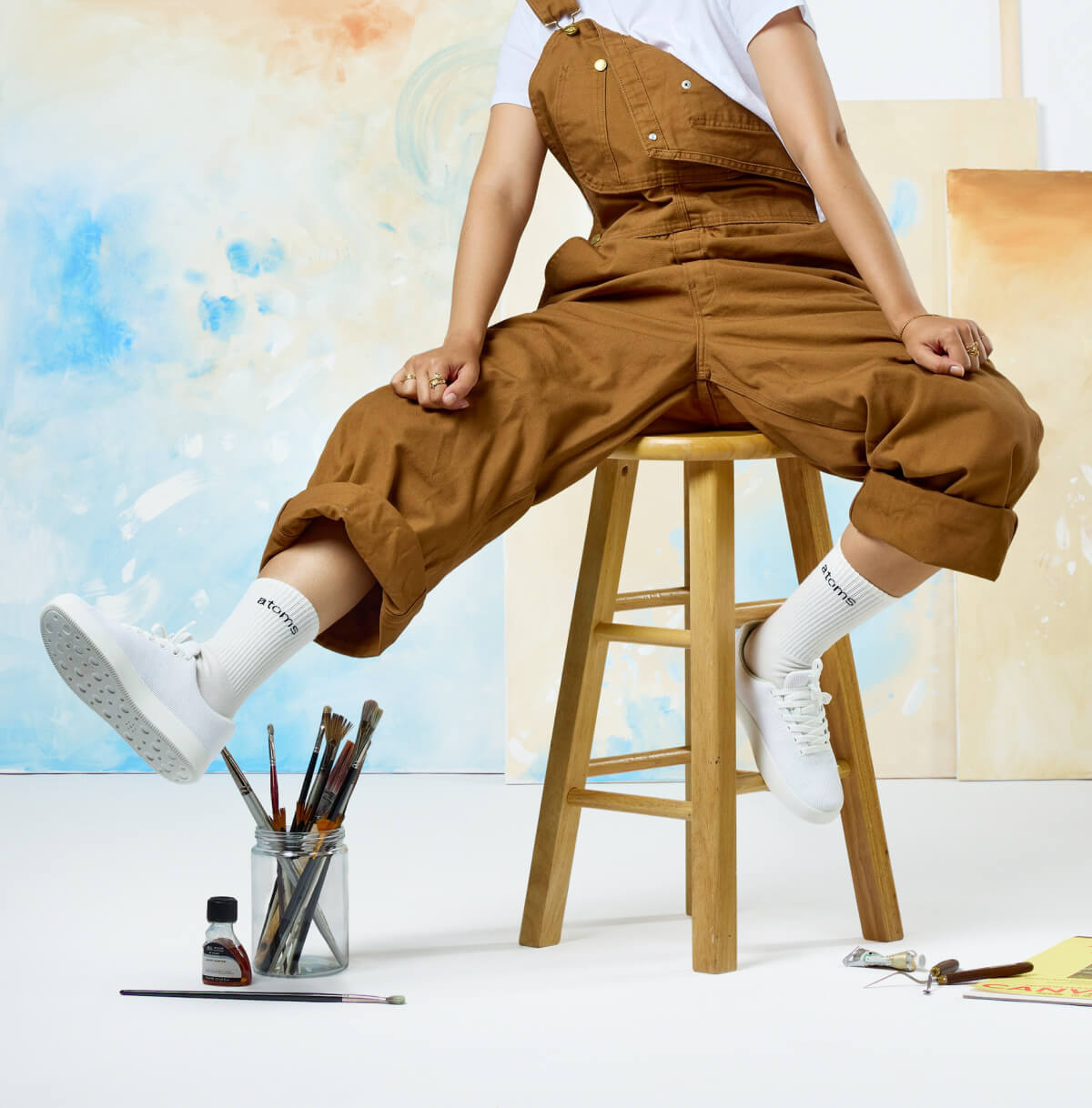 A person wearing brown overalls and white sneakers sits on a wooden stool in an art studio.