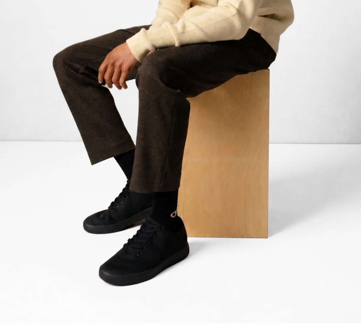 Person in beige sweater and brown pants sits on a wooden block, wearing black sneakers and socks, against a plain background.