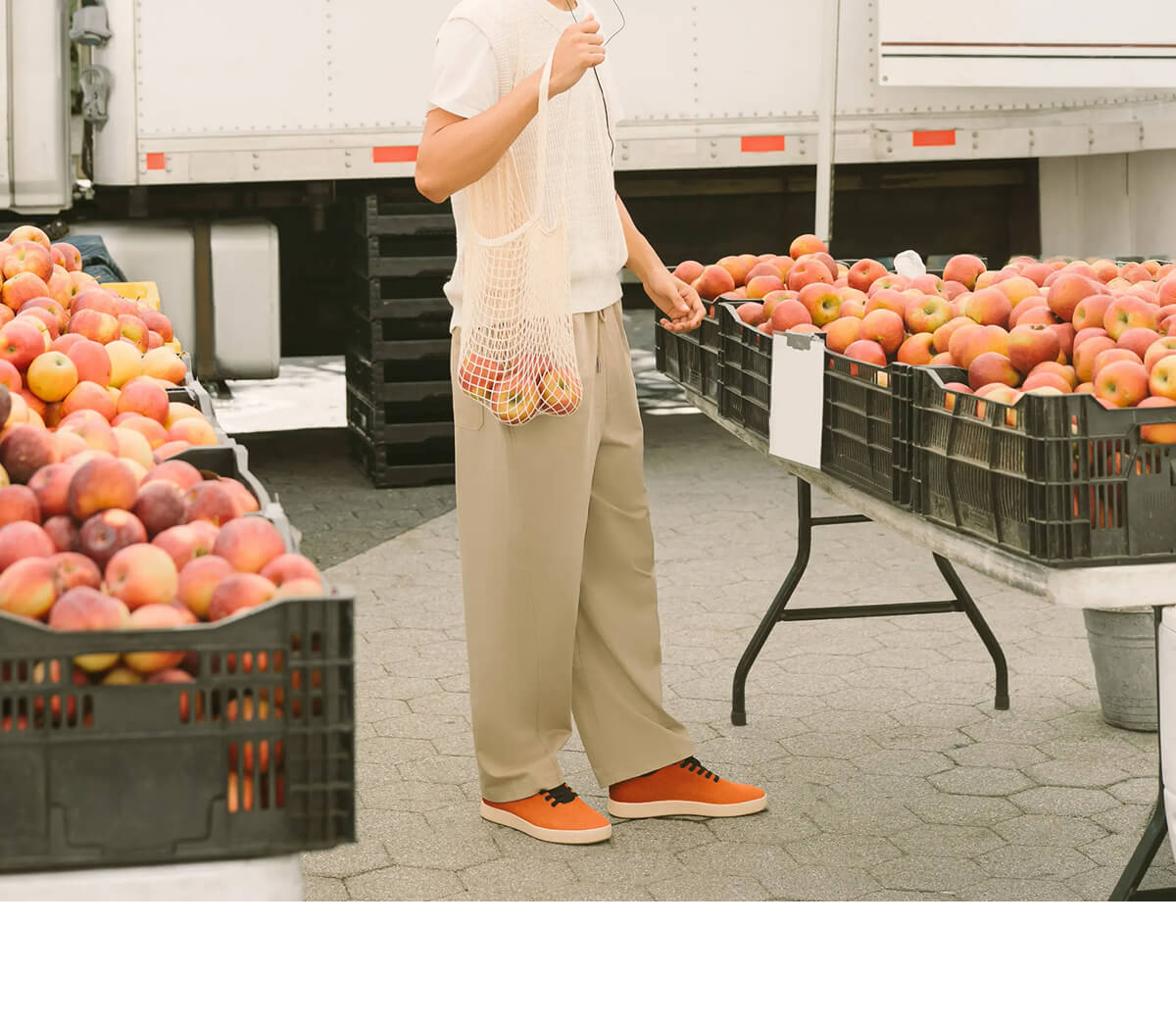 A person in a white top and beige pants holds a mesh bag of apples near crates of fruit.