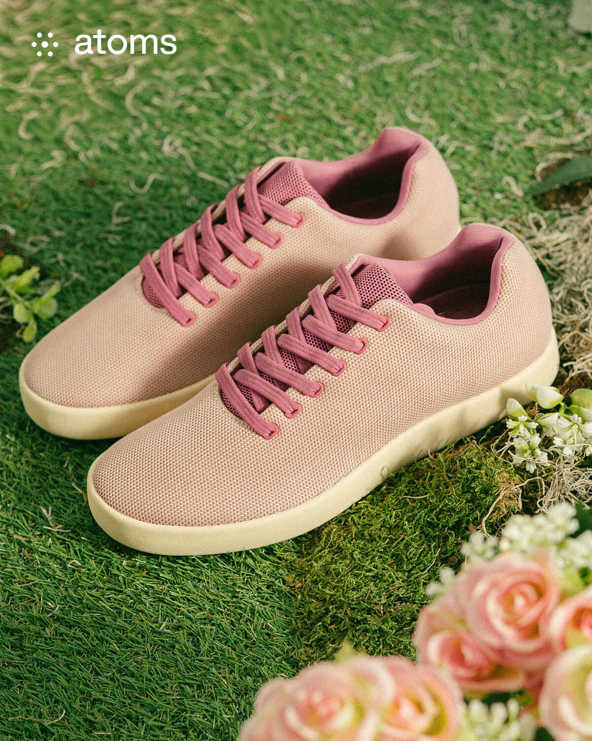 A pair of light pink sneakers with darker pink laces are placed on green grass, surrounded by soft pink flowers, creating a fresh, spring-like vibe.