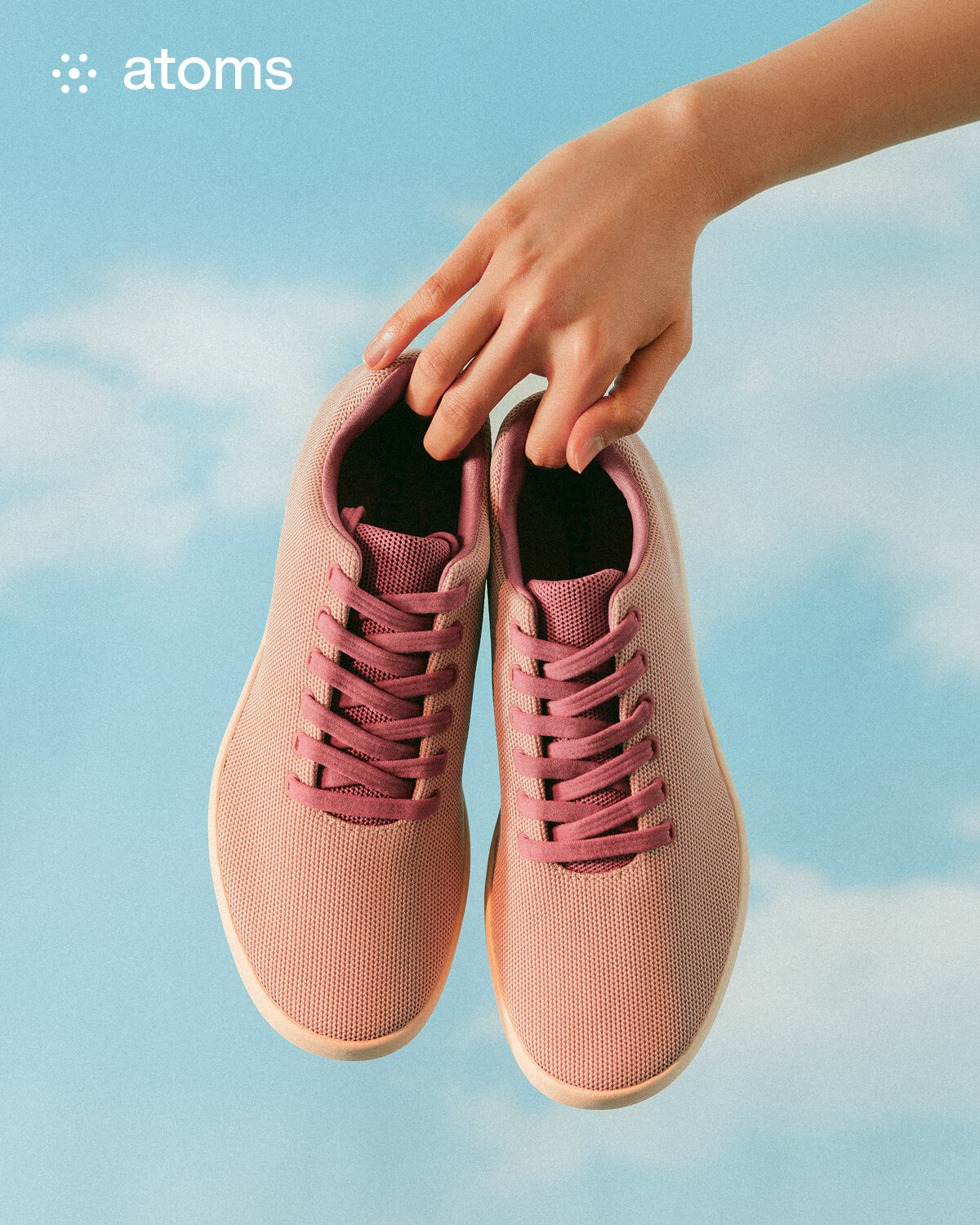 A hand holds a pair of pink sneakers with red laces against a bright blue sky with soft clouds. The mood is fresh and airy, suggesting comfort and style.