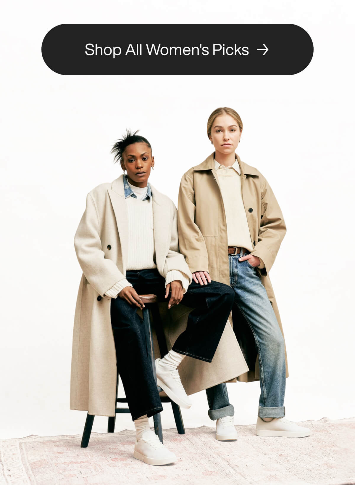 Two models wearing beige trench coats over light sweaters and jeans. One sits, the other stands, both in white sneakers. Text: "Shop All Women's Picks."