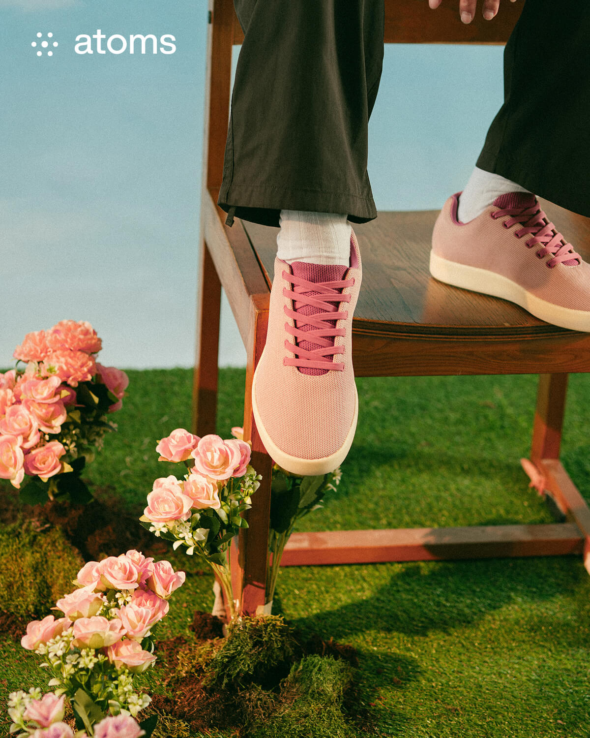 A person wearing pink sneakers sits on a wooden chair on grass, surrounded by blooming pink roses. The scene conveys a fresh and serene atmosphere. Logo text reads "atoms."