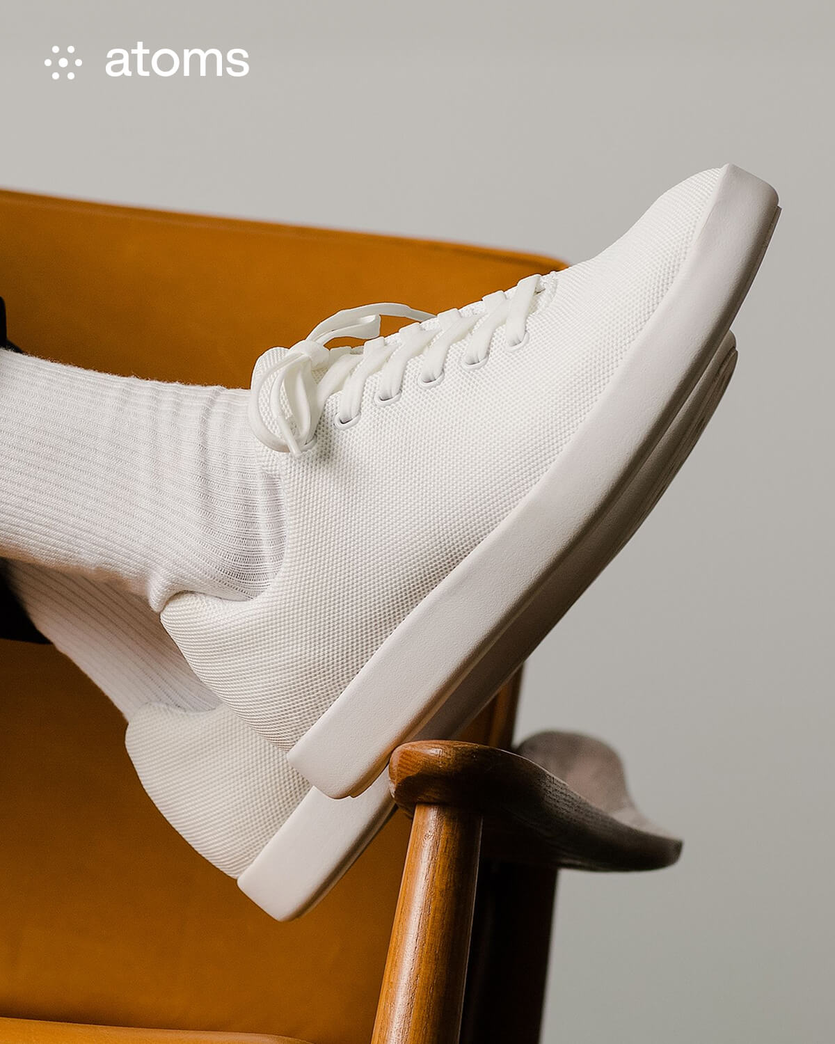 White sneakers with white laces and socks rest on the arm of a mustard-colored chair. The atmosphere is minimalist and stylish, conveying comfort.