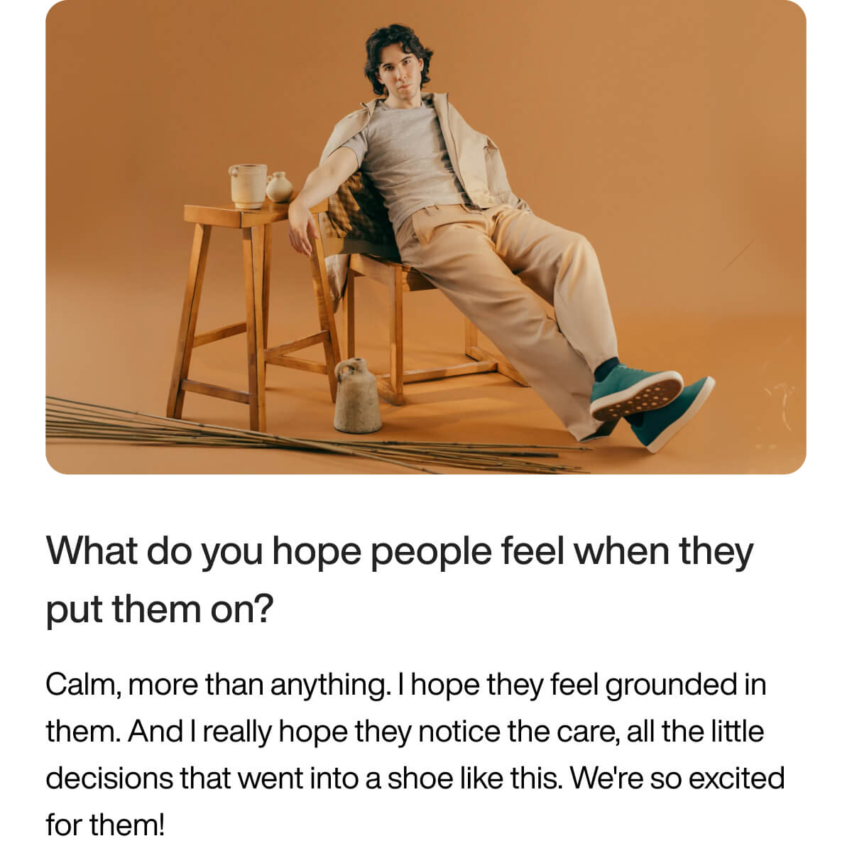 A person in casual attire sits on a wooden stool against a warm beige background, creating a calm mood. Their blue shoes stand out. There's a small wooden table nearby with ceramic items, adding a cozy touch.