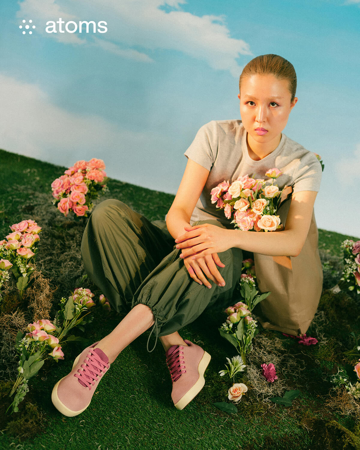 A person sits on grass, surrounded by pink flowers, holding a bouquet. They wear a gray shirt, green pants, and pink shoes. The background is a clear blue sky, conveying calmness.