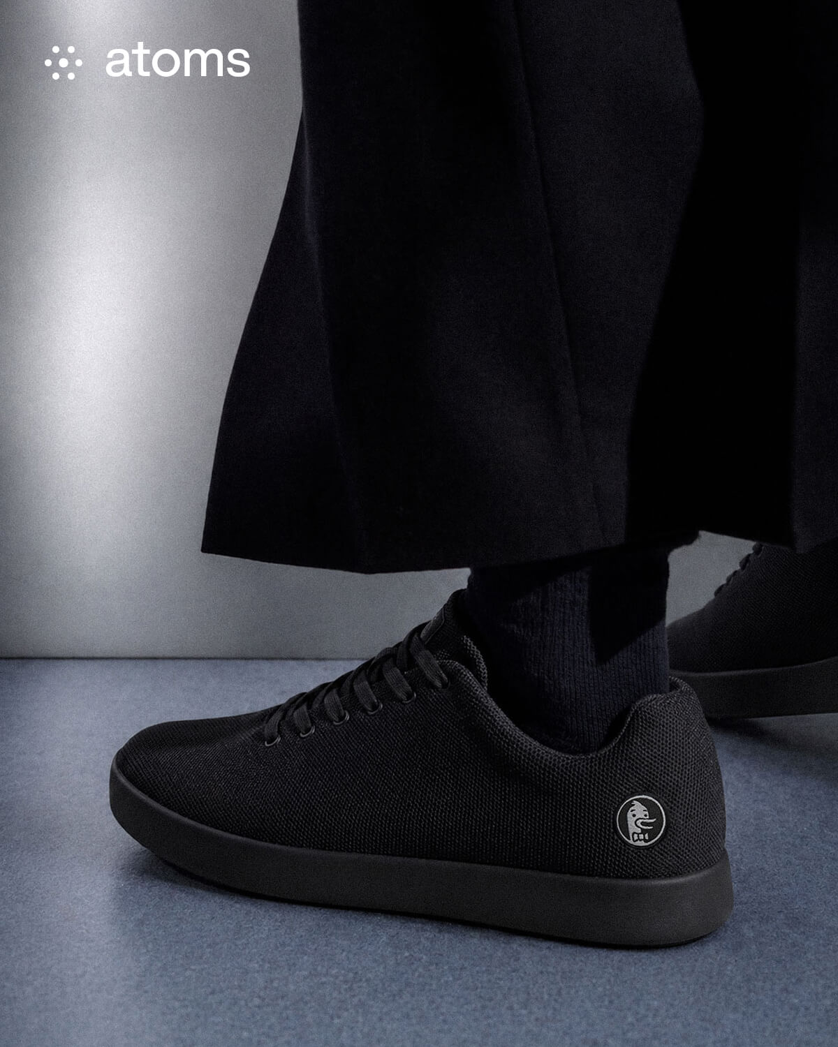 A person wearing black sneakers with a minimalist logo stands on a blue-gray floor. The backdrop is sleek and modern, conveying a stylish, refined look.